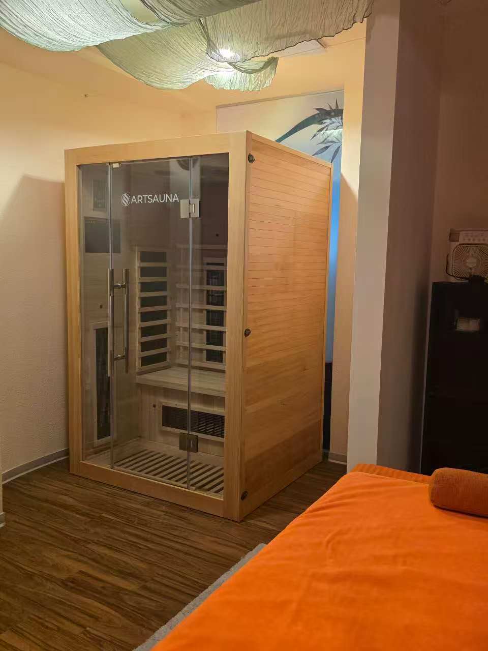 Infrarot-Kabine aus hellem Holz mit Glastür neben einer Massageliege